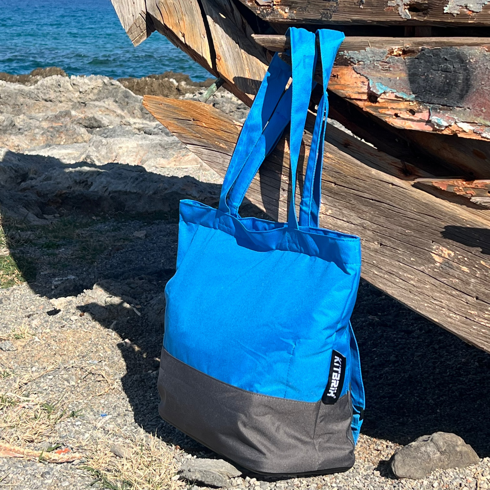 Blue and grey tote bag on a rocky beach with an old boat