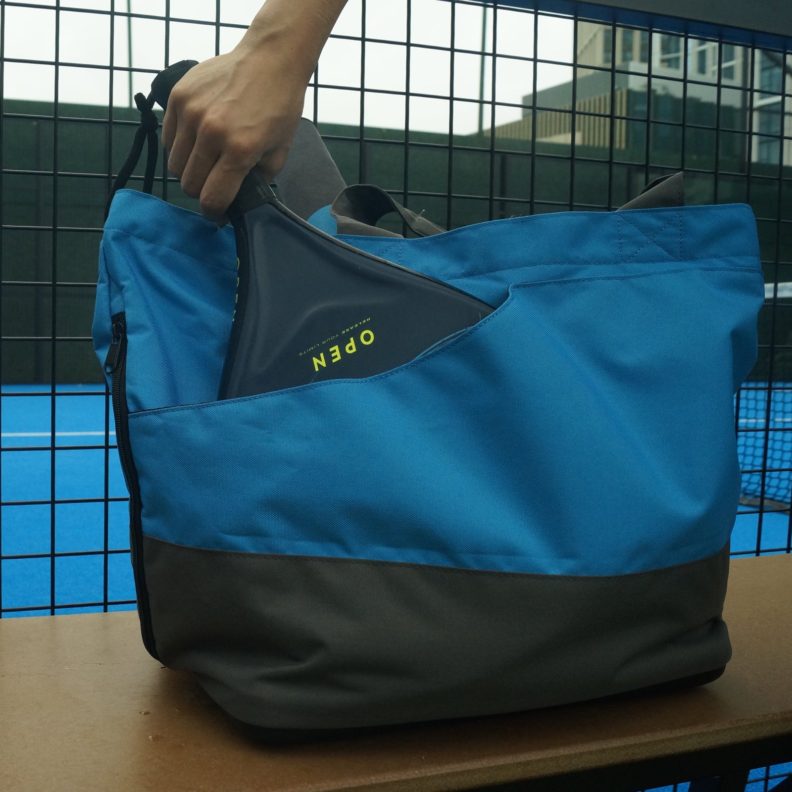 Blue and gray padel bag on a chair, with a person playing padel tennis in the background.