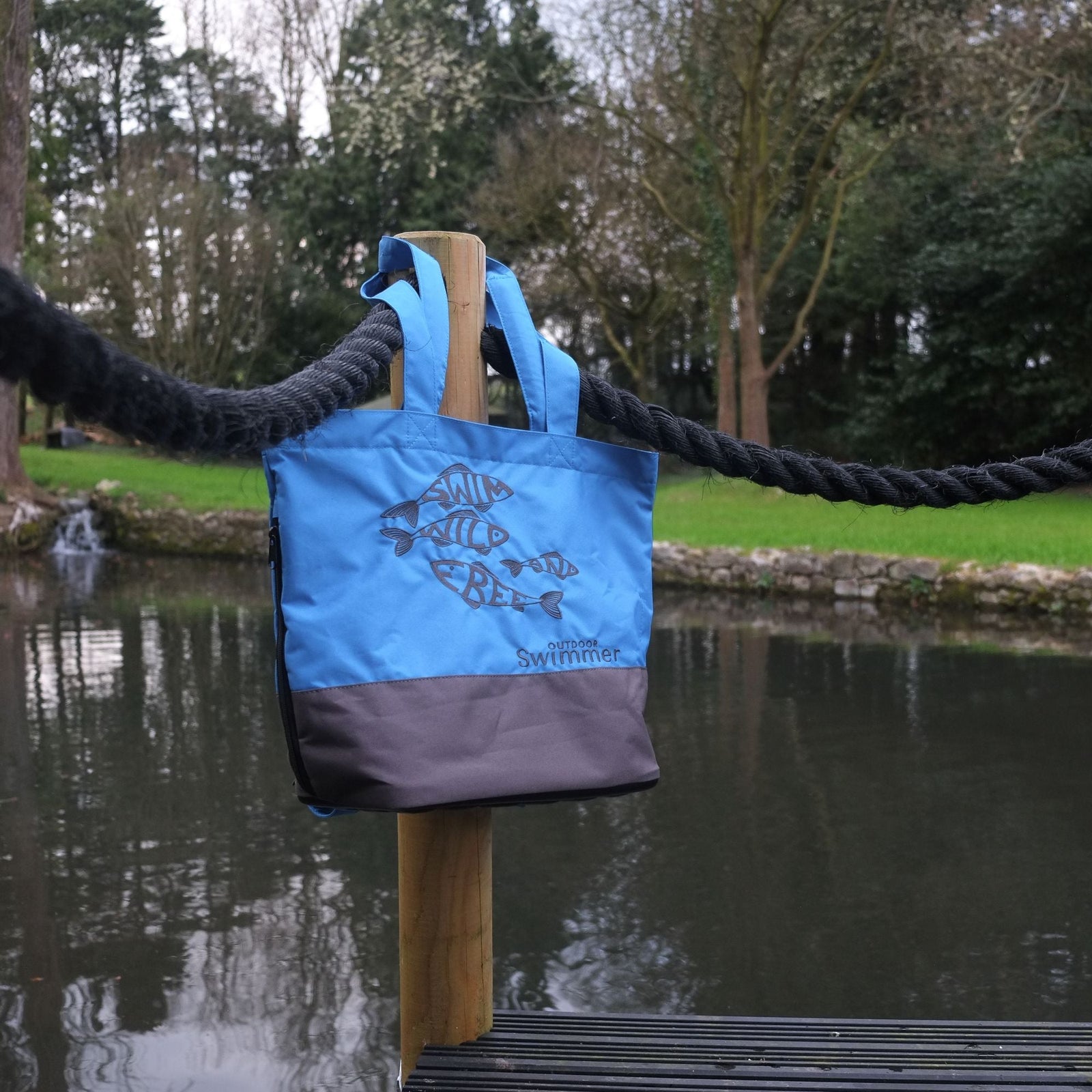 Person wearing a blue and gray shoulder bag with 'Outdoor Swimmer' branding near a body of water.