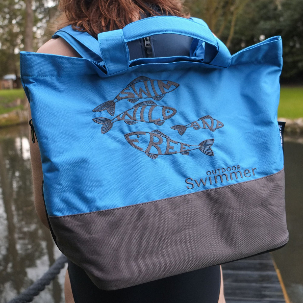 Person wearing a blue and gray shoulder bag with 'Outdoor Swimmer' branding near a body of water.