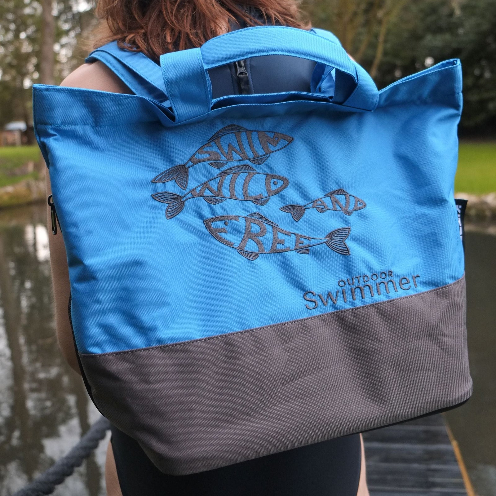 Person wearing a blue and gray shoulder bag with 'Outdoor Swimmer' branding near a body of water.