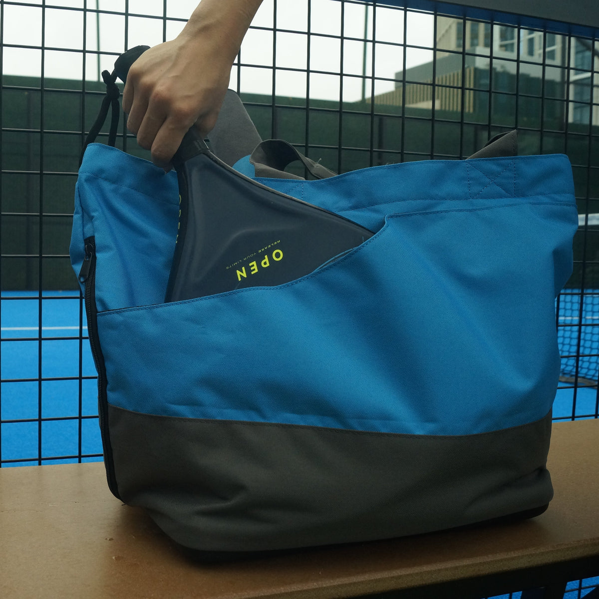 Blue and gray padel bag on a chair, with a person playing padel tennis in the background.