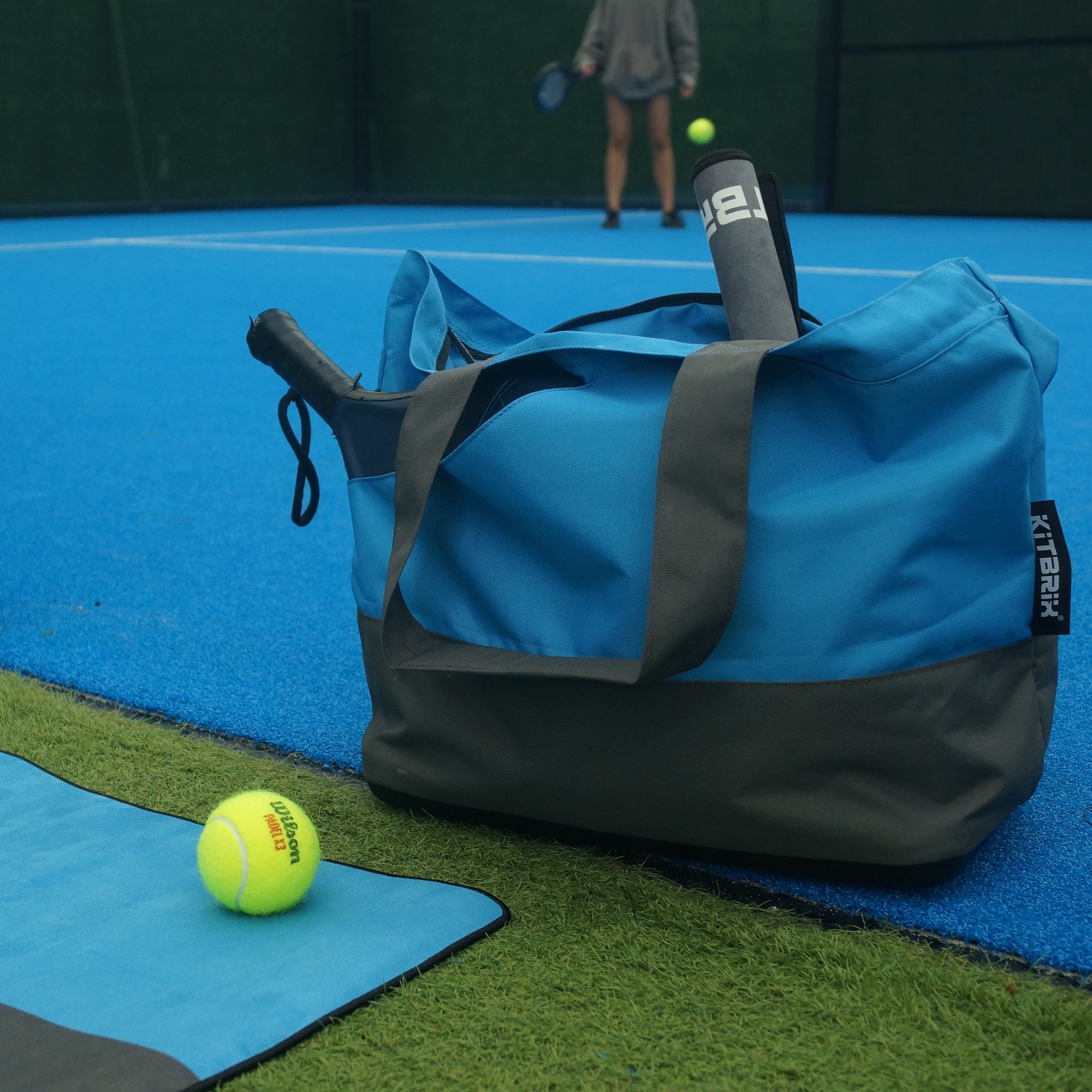 Blue and gray tennies tote bag with a tennis ball  and mat on tennis court
