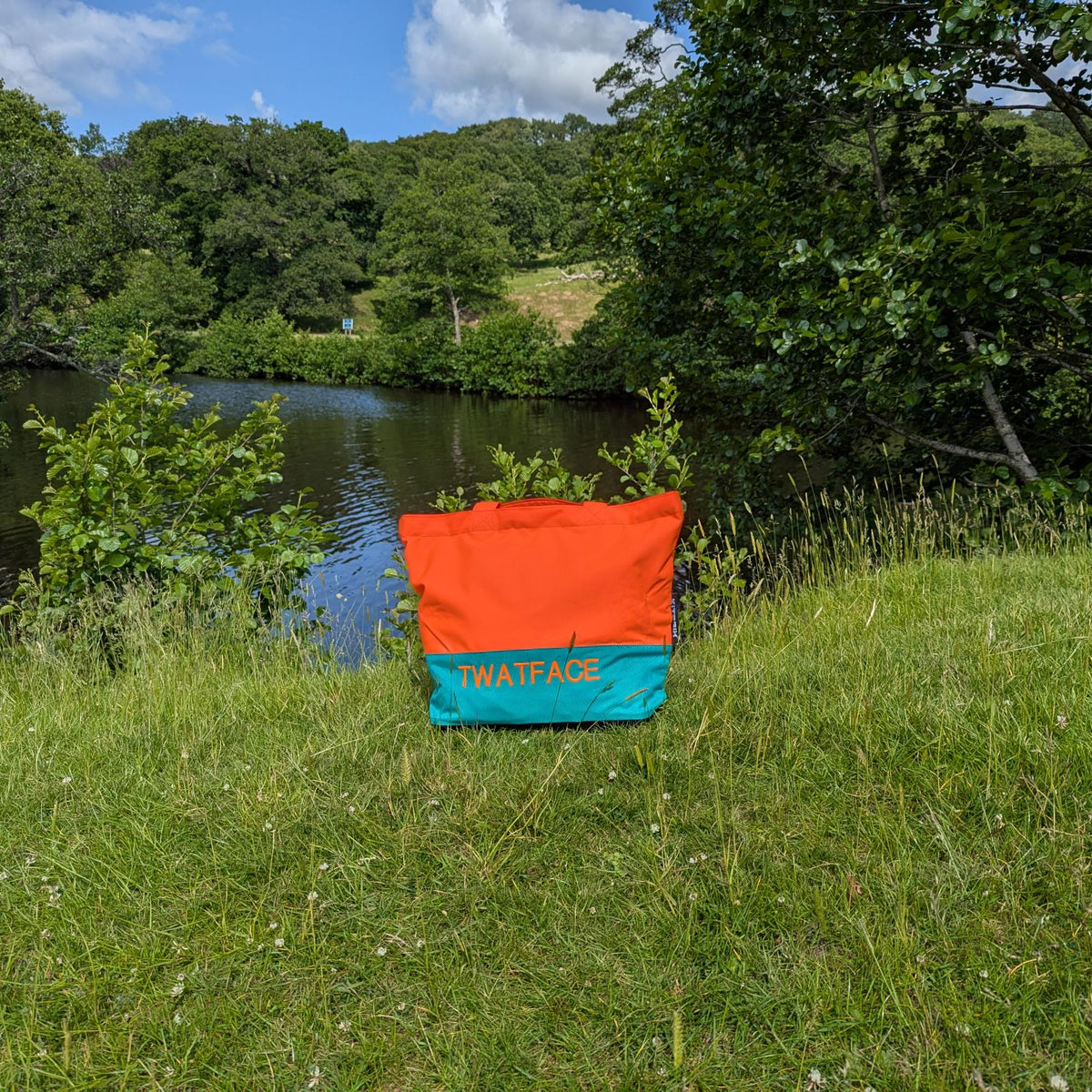 Orange and blue bag with 'WATFACE' customised logo on grass near a pond and trees